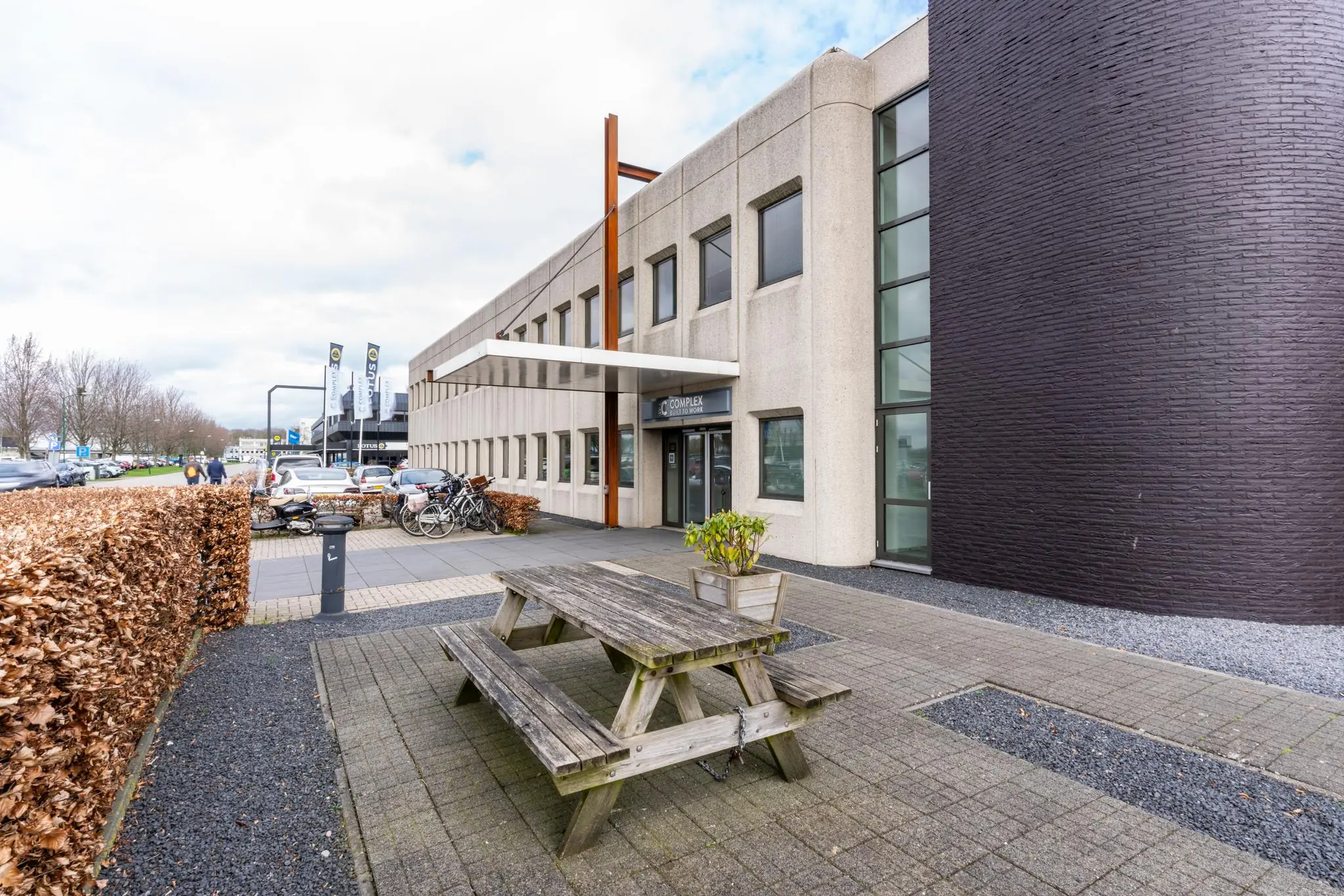 Vooraanzicht van een modern kantoorgebouw aan de Meidoornkade met fietsenrek, parkeerplaatsen en een picknicktafel op de voorgrond.
