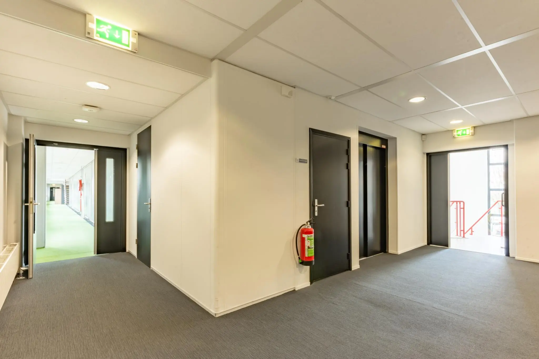 Binnenhal met zwarte deuren, een lift, vluchtwegaanduiding en een brandblusser in een gebouw aan de Meidoornkade.