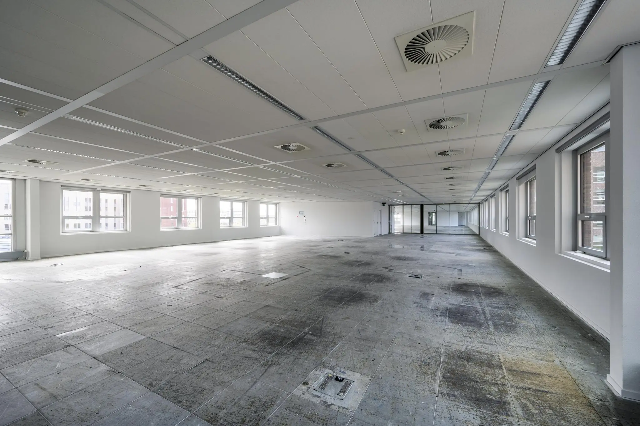 Leeg kantoorpand aan de Orteliuslaan met betonnen vloer, systeemplafond en grote ramen aan de rechterzijde.