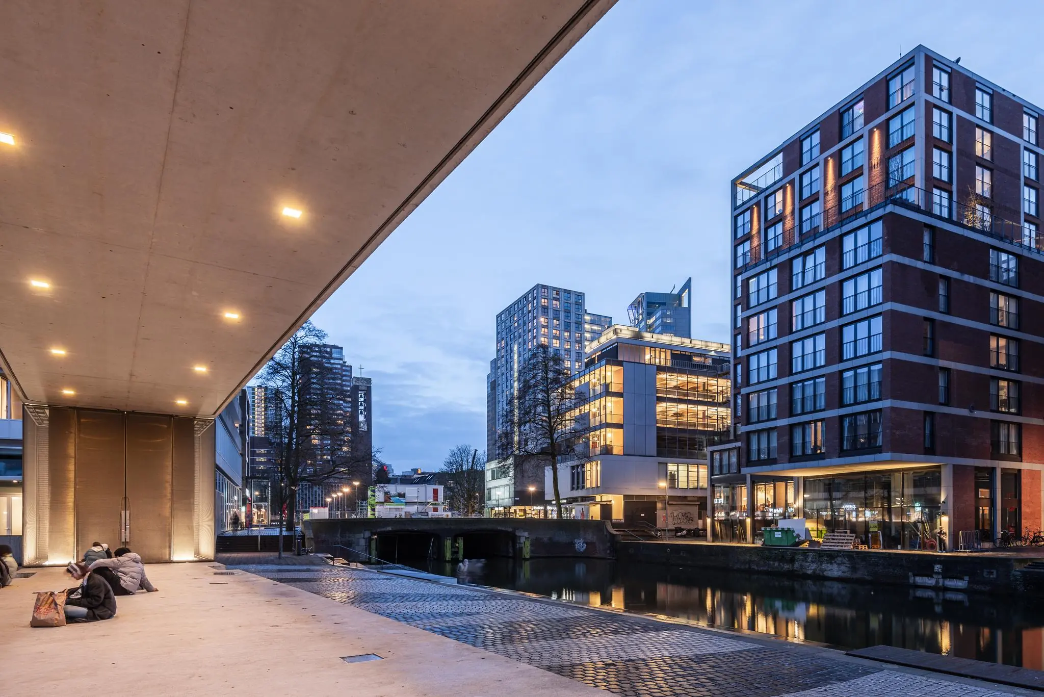 Avondzicht op de Vlasmarkt met moderne gebouwen, een kanaal en enkele zittende mensen onder een overkapping.