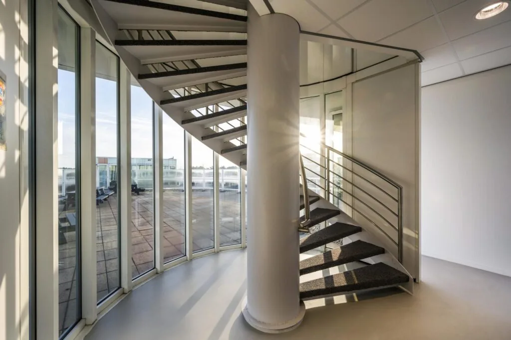 Moderne, ronde wenteltrap bij Paalbergweg met glazen wand en uitzicht op een dakterras.