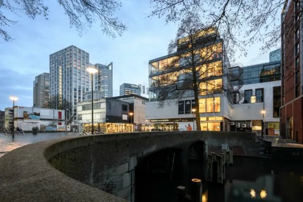 Gezicht op de Vlasmarkt in Rotterdam met moderne hoogbouw, winkels en een stenen brug over het water bij schemering.