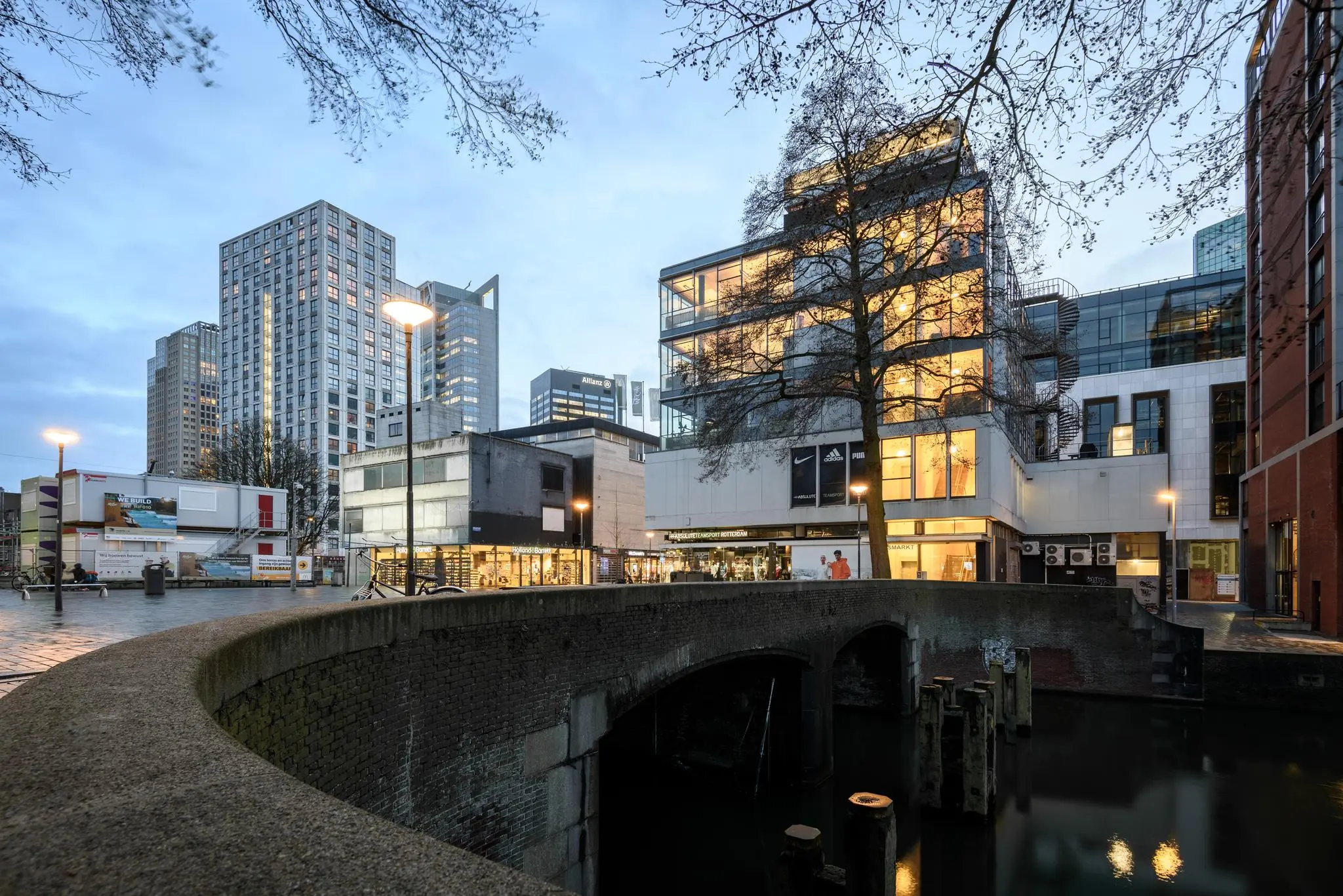 Gezicht op de Vlasmarkt in Rotterdam met moderne hoogbouw, winkels en een stenen brug over het water bij schemering.