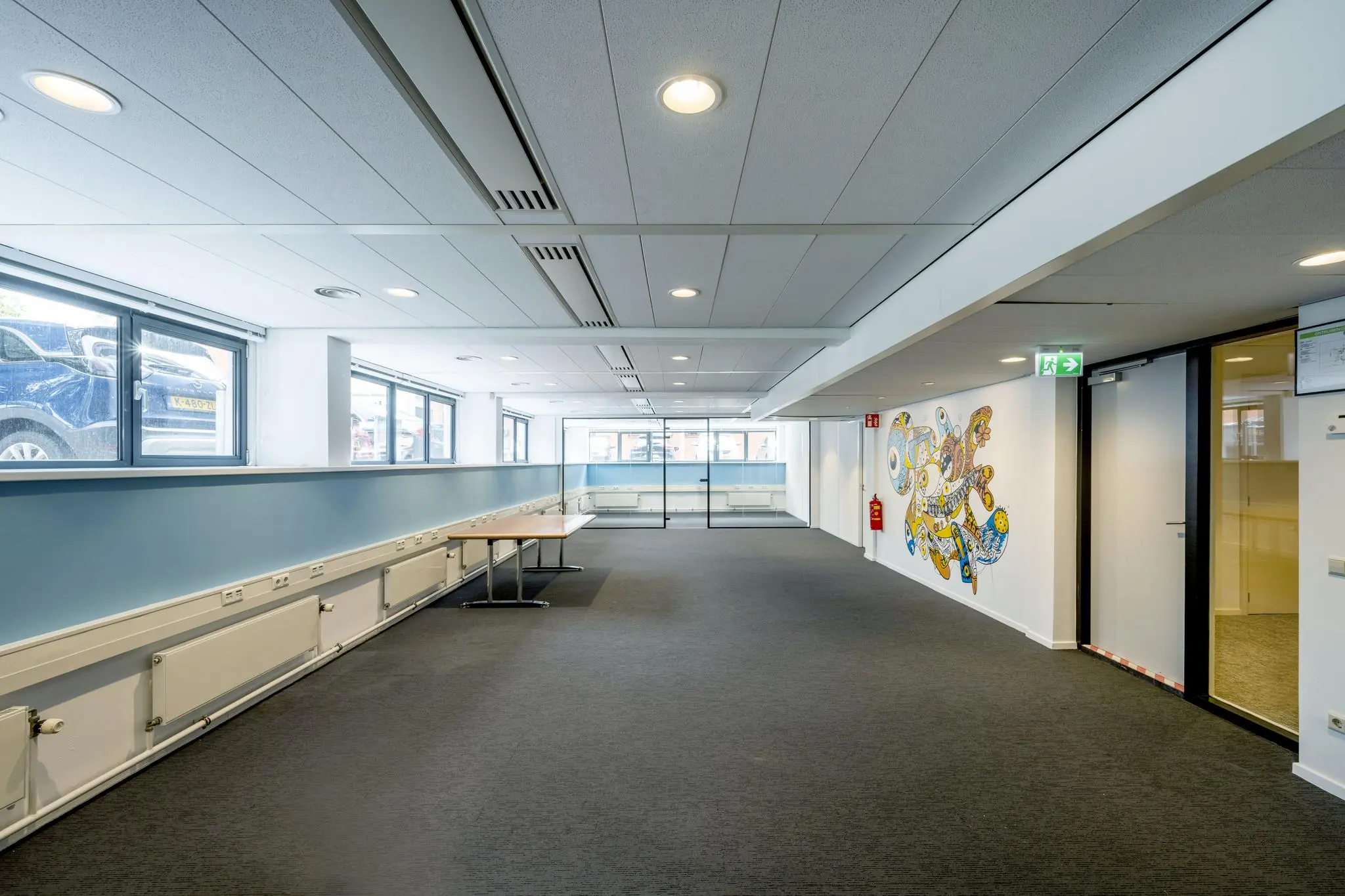 Lichte kantoorruimte met tapijtvloer, glazen vergaderruimte, muurschildering en ramen met uitzicht op geparkeerde auto's.