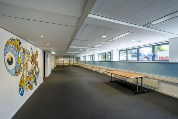 Moderne kantoorruimte met kunstzinnige muurschildering aan de linkerwand en ramen met uitzicht op geparkeerde auto's rechts.