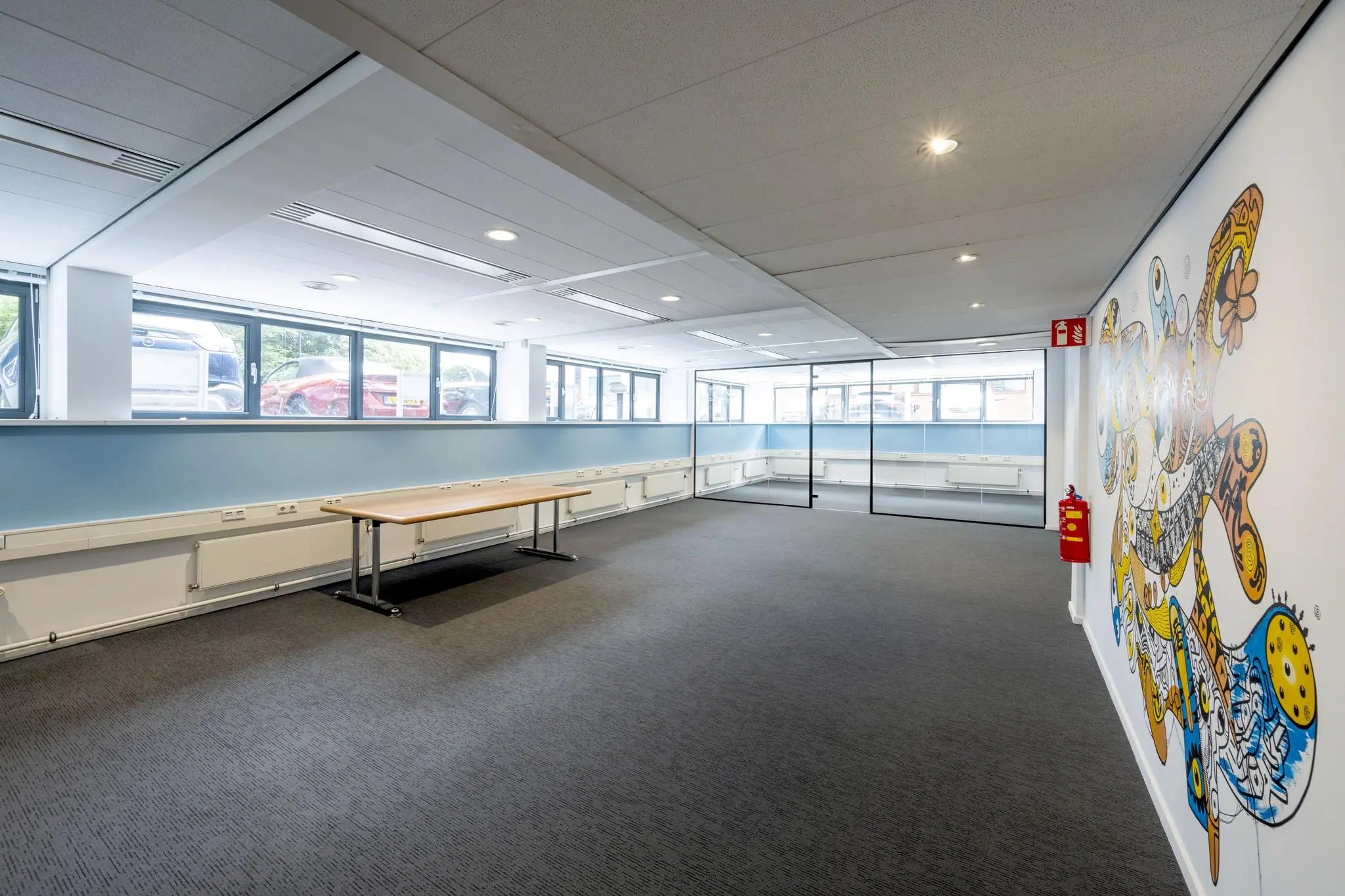 Ruime, moderne kantoorruimte met grote ramen, glazen wanden en een kleurrijke muurschildering aan de rechterkant.
