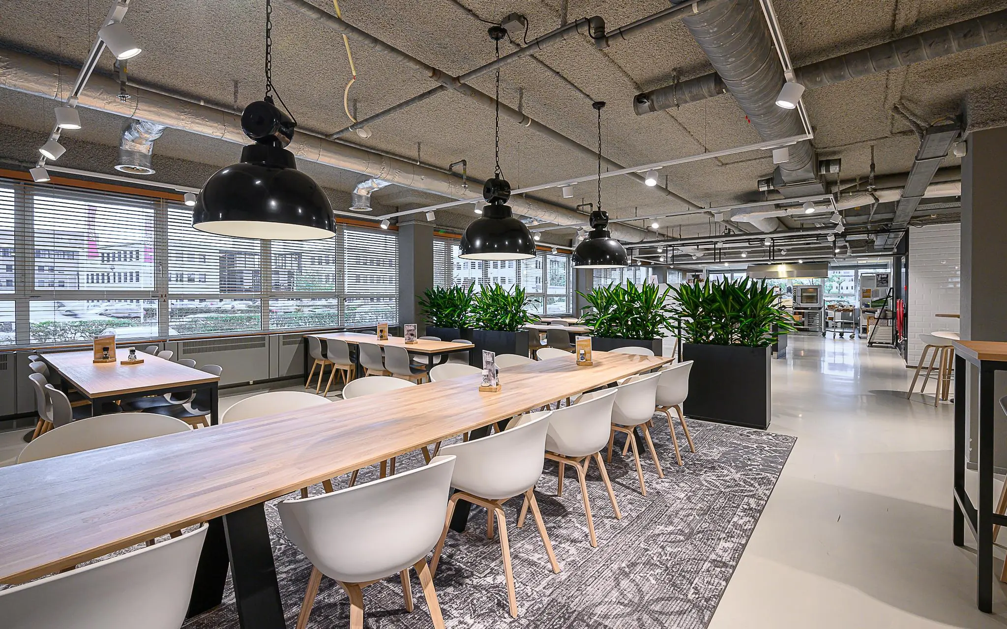 Modern ingerichte bedrijfsrestaurant aan de Rivium 1e Straat met lange houten tafels, witte stoelen, hanglampen en grote plantenbakken.