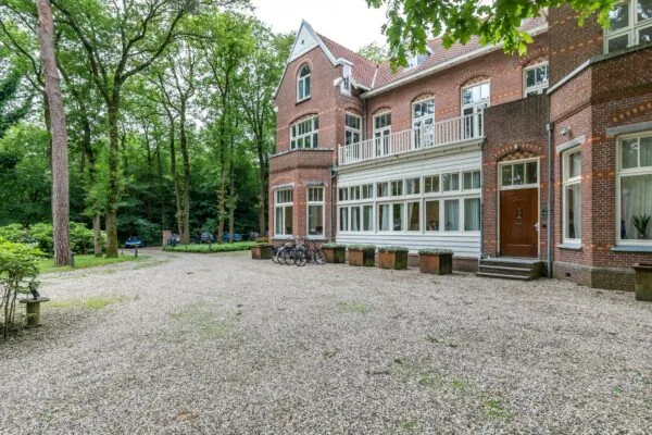 Vooraanzicht van een statig herenhuis aan de Amersfoortsestraat, omgeven door bomen en geparkeerde fietsen op een grindterrein.