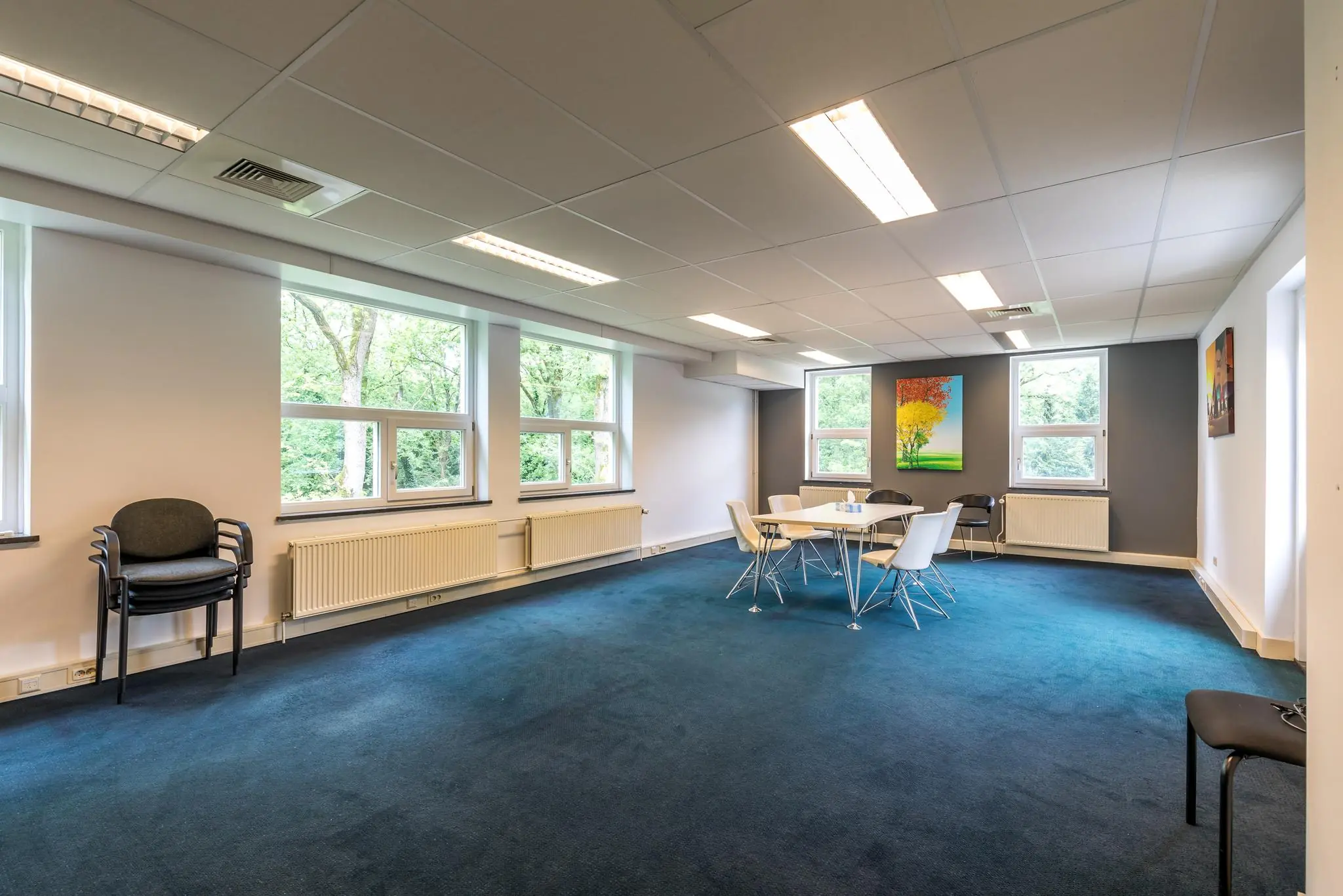 Vergaderruimte aan de Amersfoortsestraat met blauwe vloerbedekking, grote ramen en een ronde tafel met stoelen.