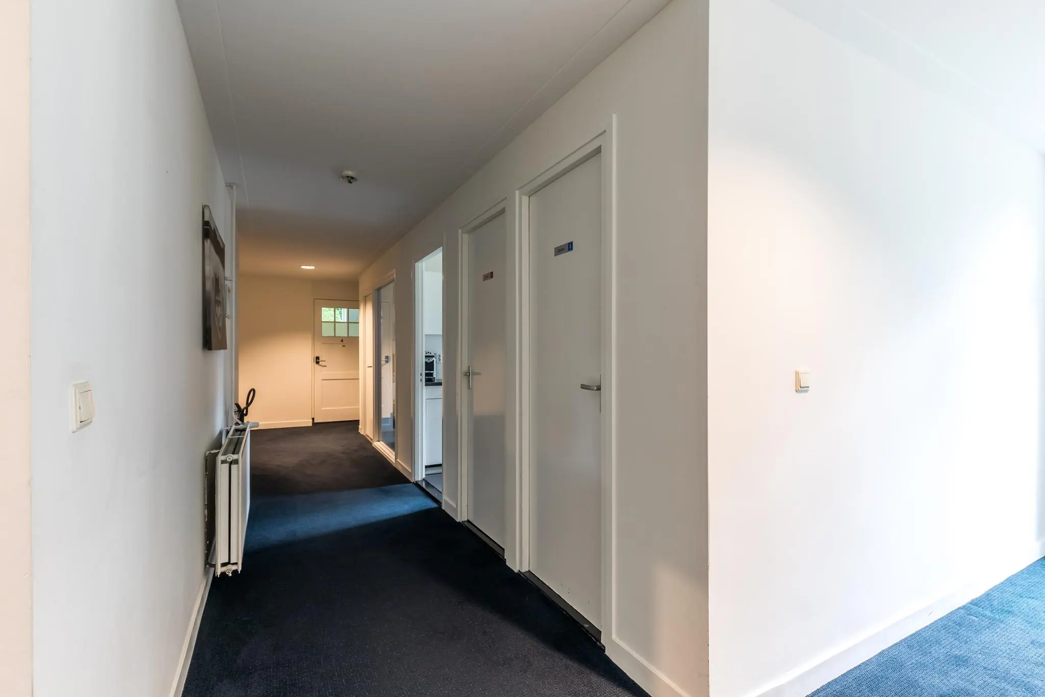 Verlichte gang in een gebouw aan de Amersfoortsestraat met meerdere deuren en donkerblauwe vloerbedekking.