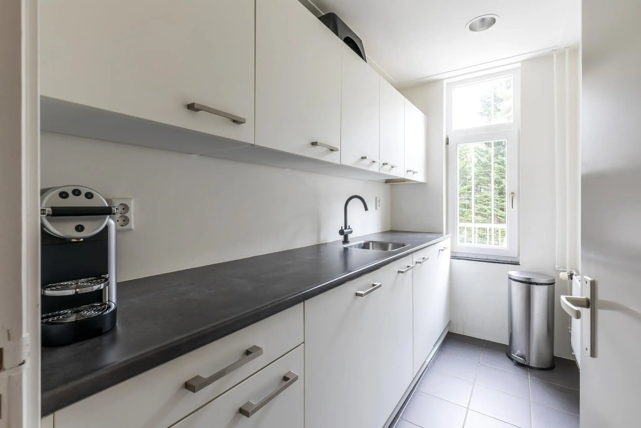 Smalle, moderne keuken aan de Amersfoortsestraat met witte kasten, zwarte werkblad, koffiemachine en een raam met uitzicht op groen.