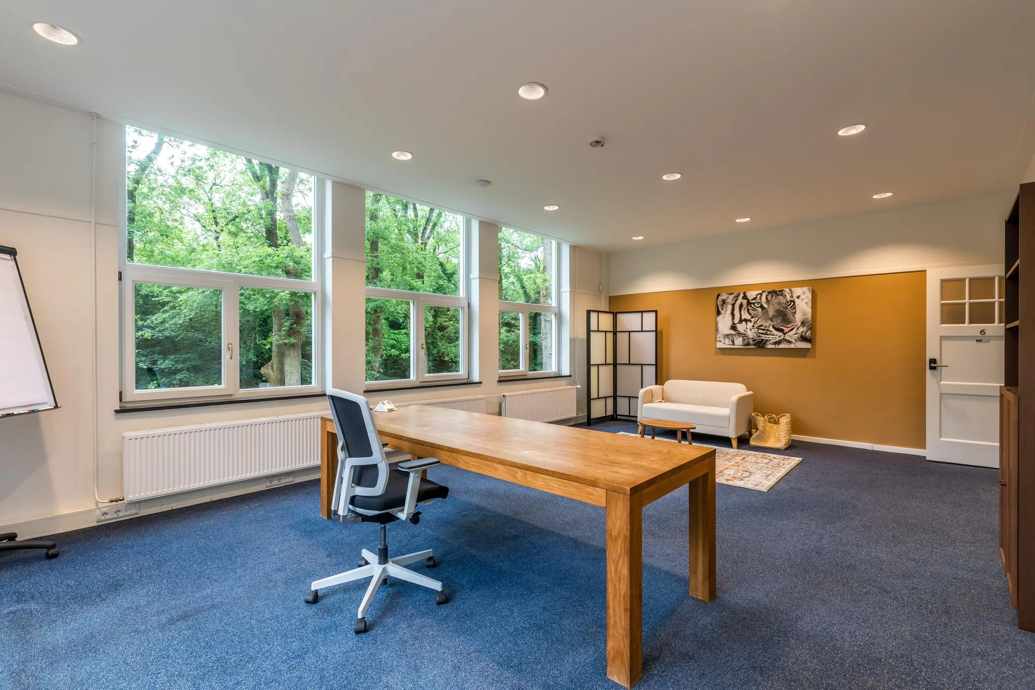 Modern kantoor met houten bureau, grote ramen met uitzicht op groen en een zithoek met tijgerkunst aan de muur.