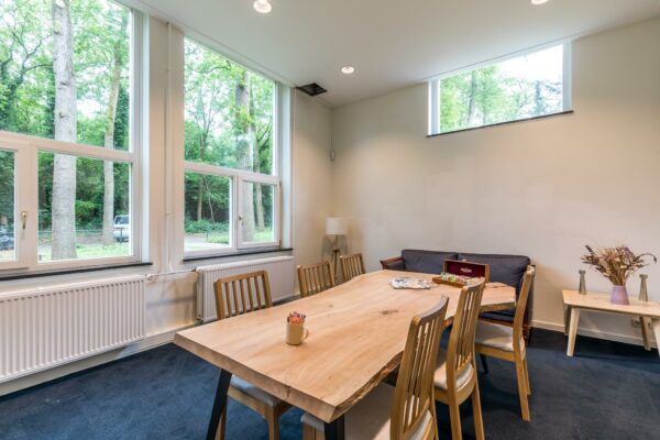 Vergaderruimte met houten tafel, zes stoelen en grote ramen met uitzicht op een bosrijke omgeving aan de Amersfoortsestraat.