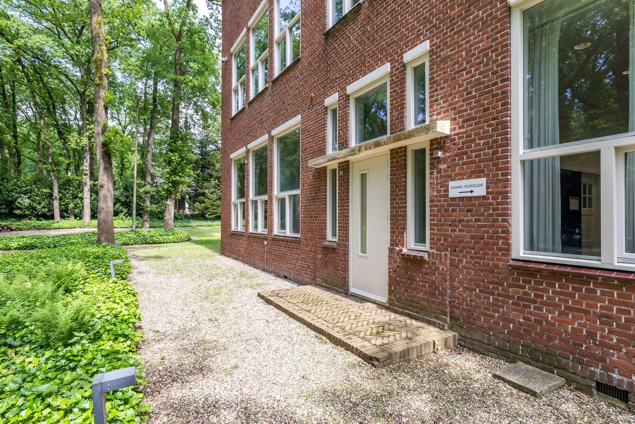 Vooraanzicht van een bakstenen gebouw aan de Amersfoortsestraat met een witte deur en omgeven door een groen bosperceel.
