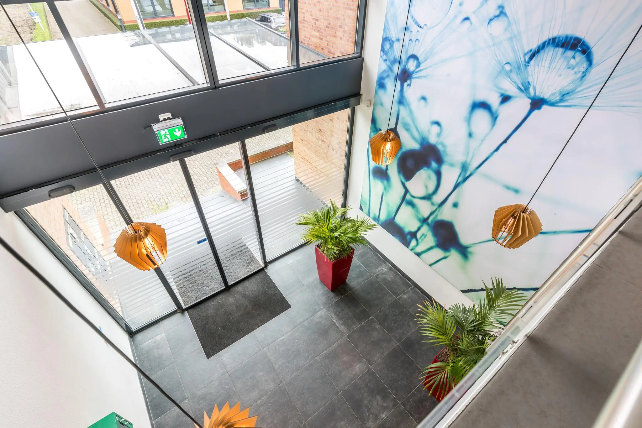 Moderne entreehal van een kantoorpand aan de Coltbaan met glazen schuifdeuren, grote wandprint van bloemen, hanglampen en planten in rode potten.
