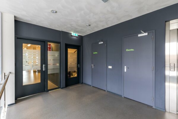 Hal met toegang tot toiletten gemarkeerd als "Ladies" en "Gents", een lift en deuren naar kantoren aan de Coltbaan.