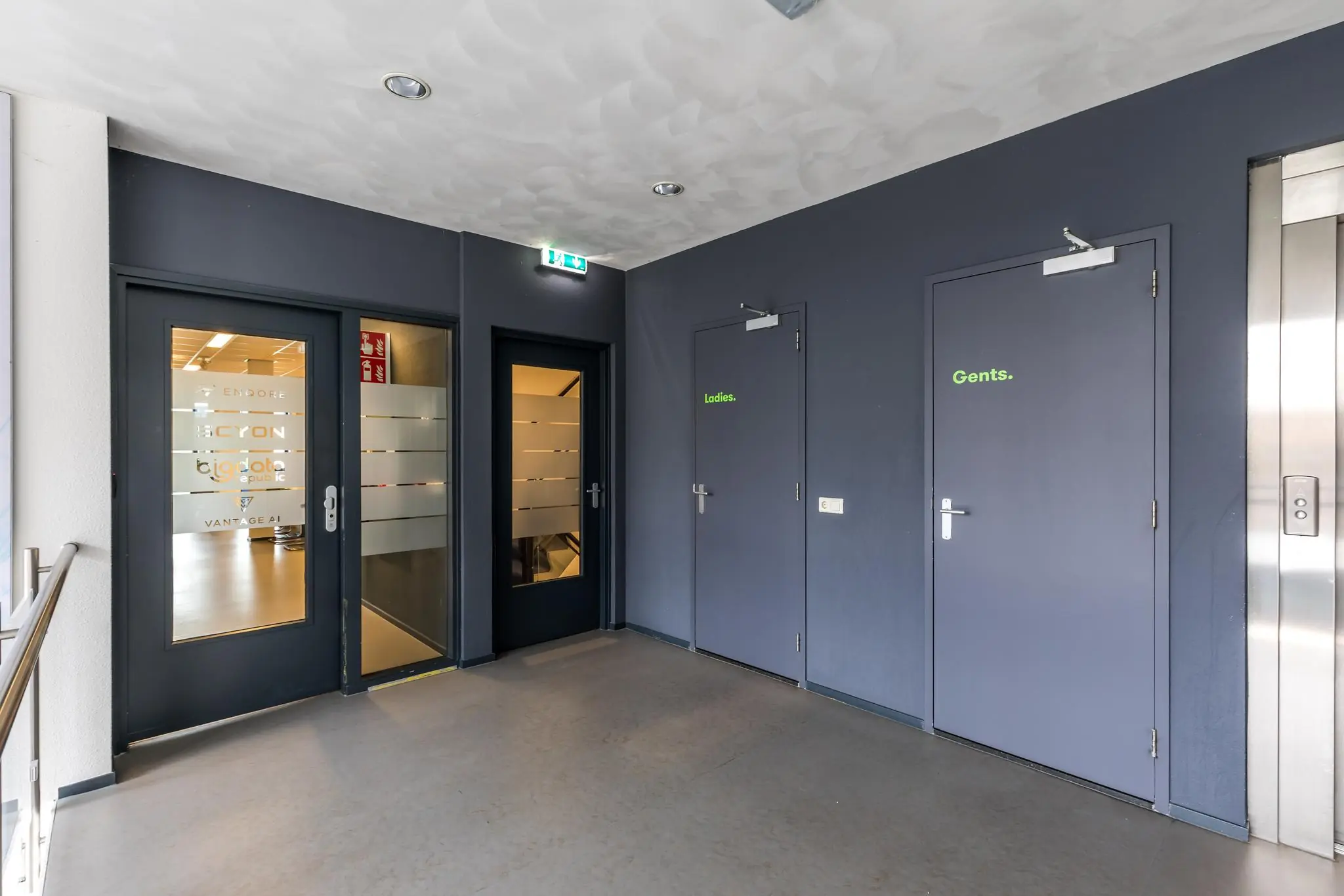 Hal met toegang tot toiletten gemarkeerd als "Ladies" en "Gents", een lift en deuren naar kantoren aan de Coltbaan.