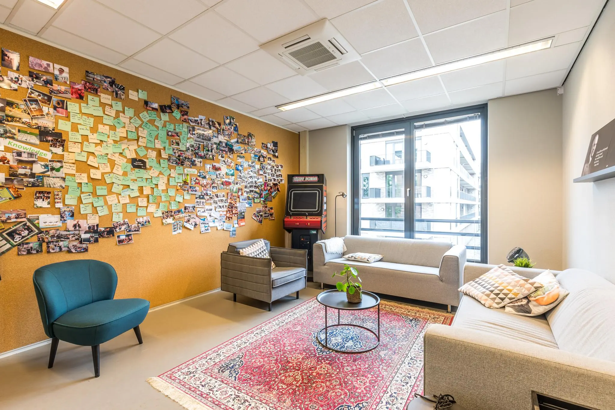 Een moderne kantoorlounge met zithoeken, een kleurrijk prikbord vol foto's en post-its, een retro arcadekast en grote ramen met uitzicht op gebouwen.