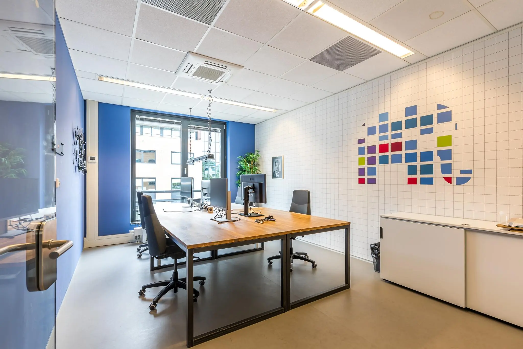 Moderne kantoorruimte aan de Coltbaan met houten bureaus, ergonomische stoelen en kleurrijke wanddecoratie op een witte tegelmuur.