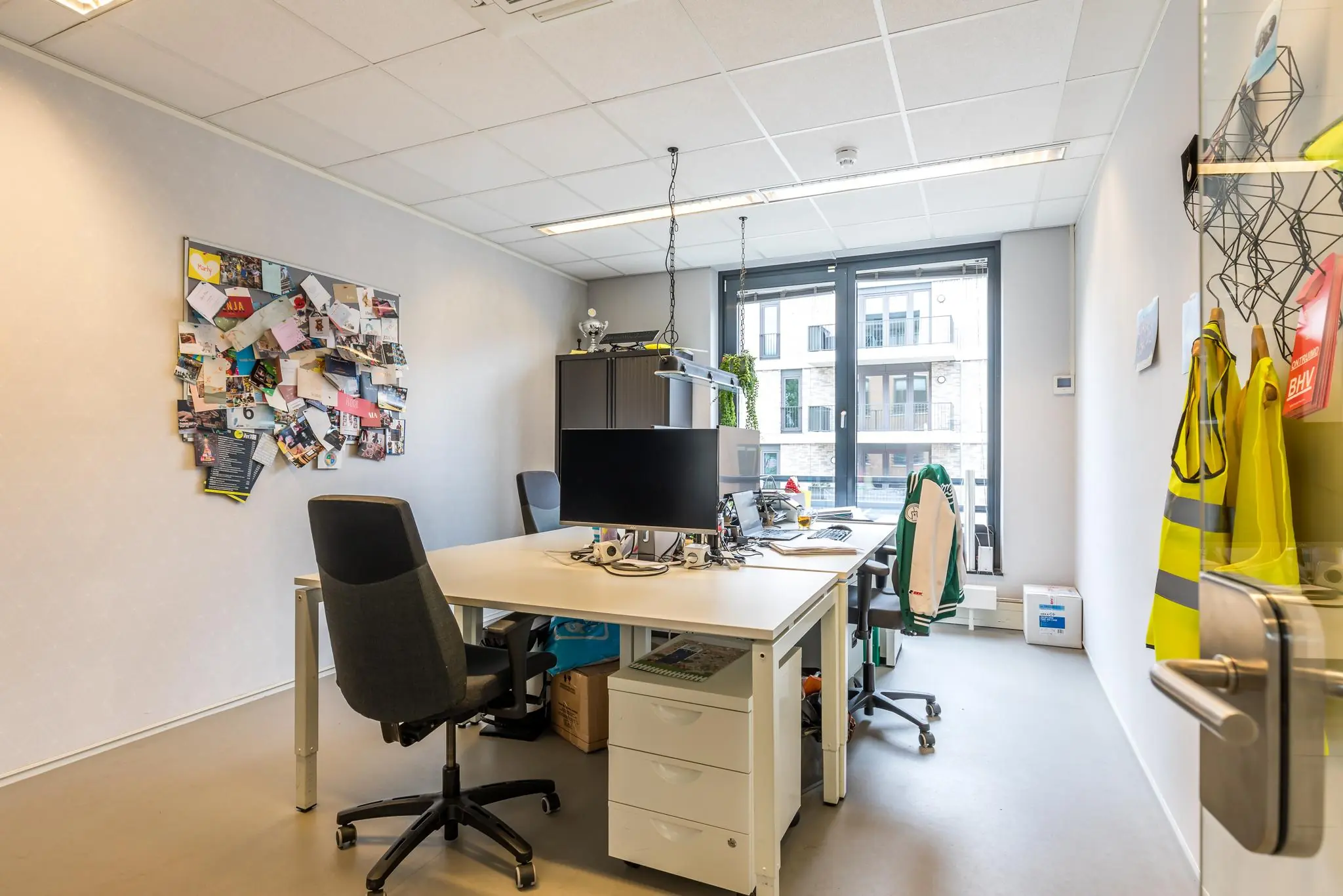 Klein en licht kantoor met twee bureaus, computerschermen, een prikbord vol foto's en kaarten, en zicht op een modern appartementencomplex.