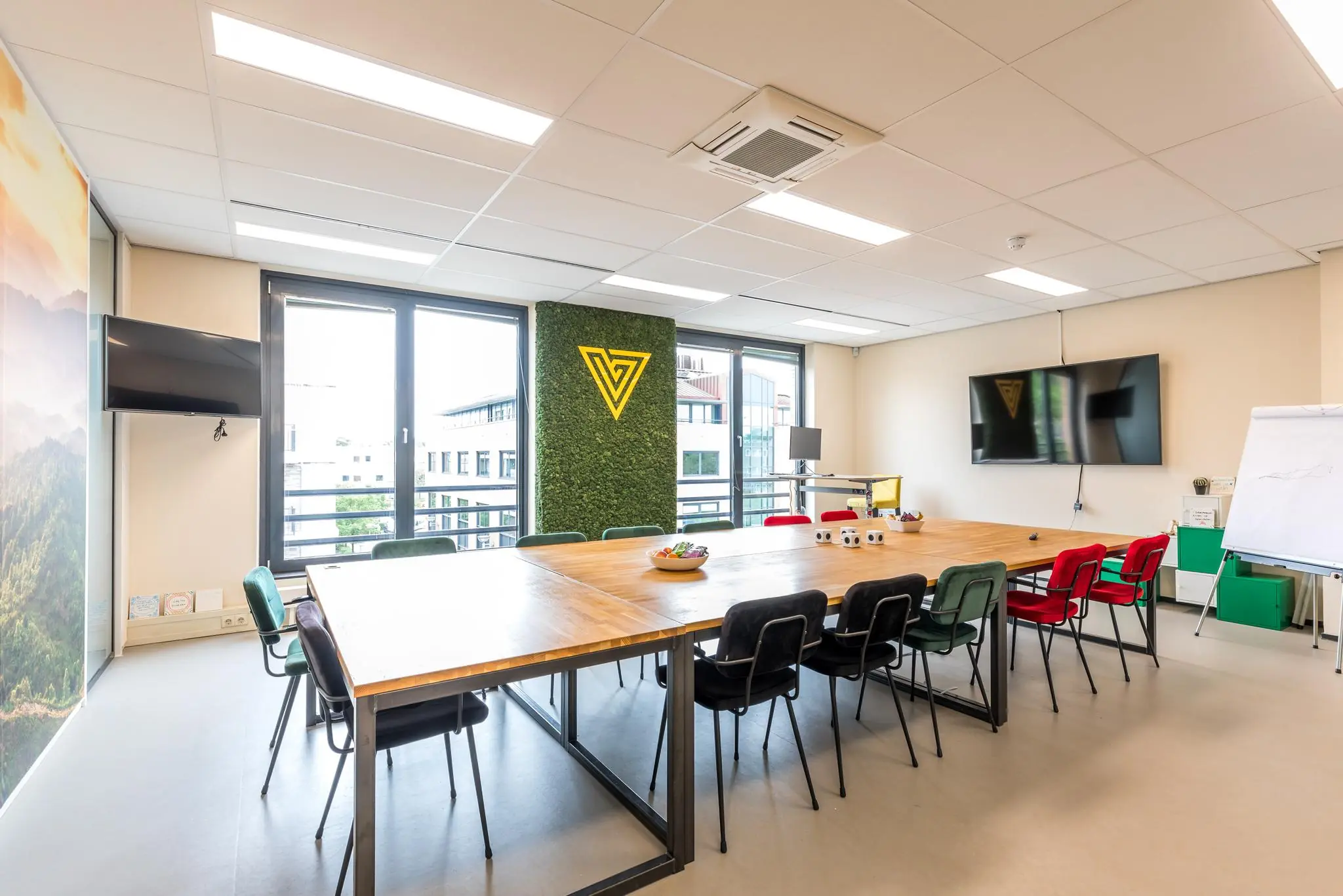 Moderne vergaderruimte aan de Coltbaan met grote houten tafel, kleurrijke stoelen, twee schermen en een groene wand met geel logo.