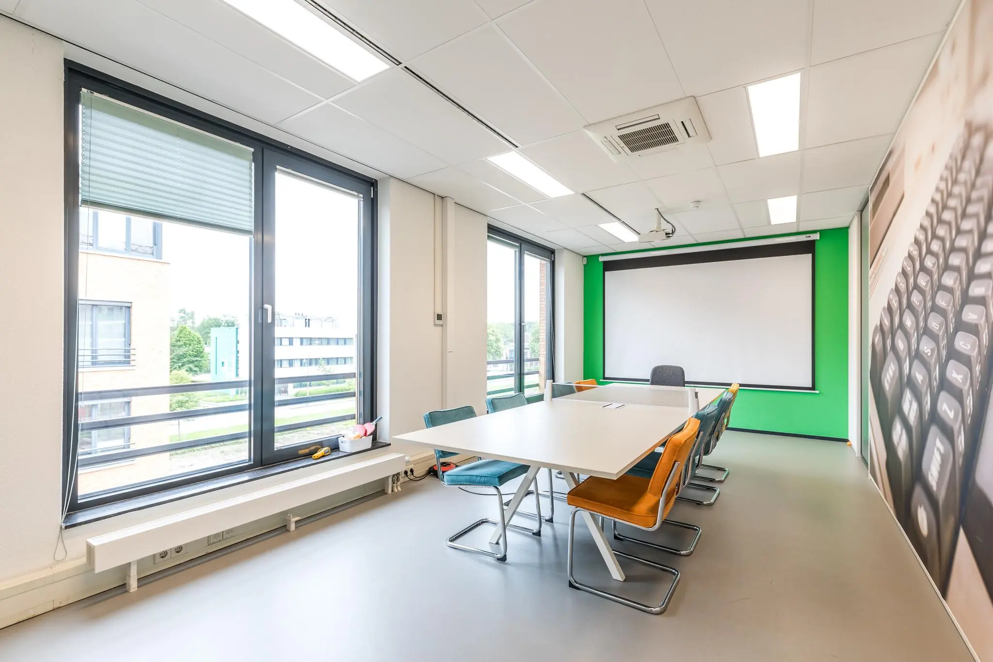 Vergaderruimte aan de Coltbaan met grote ramen, een witte tafel, kleurrijke stoelen en een projectiescherm.