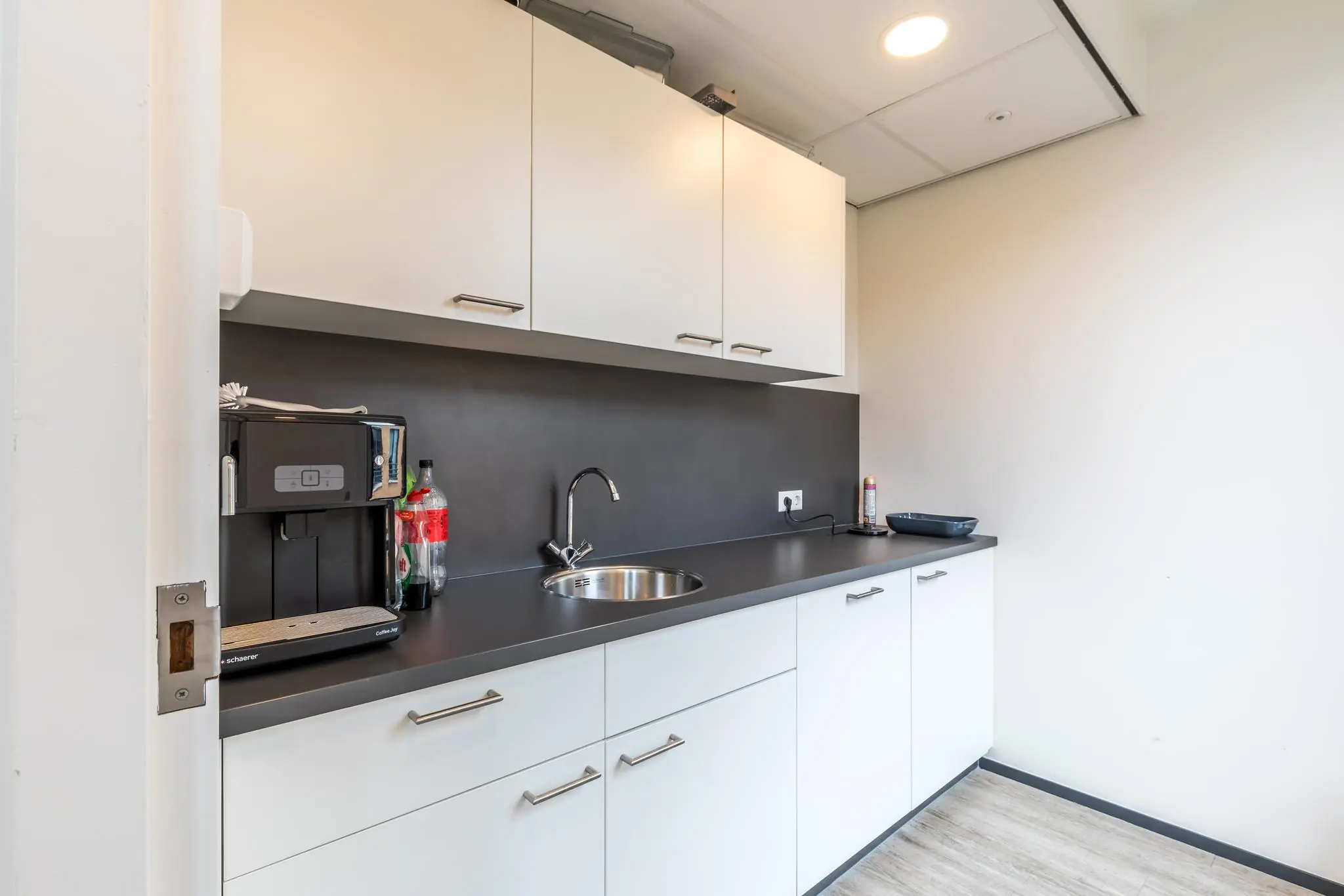 Moderne pantry met witte kasten, een koffiezetapparaat, spoelbak en zwarte werkblad in een kantooromgeving aan de Coltbaan.