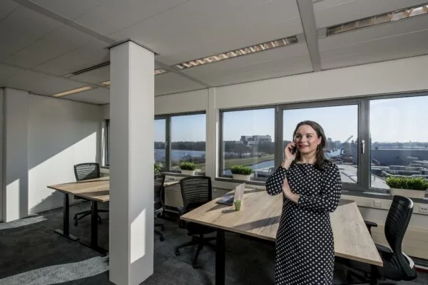 Vrouw in een modern kantoor aan de Vanadiumweg, telefonerend bij een bureau met uitzicht op een industrieterrein.