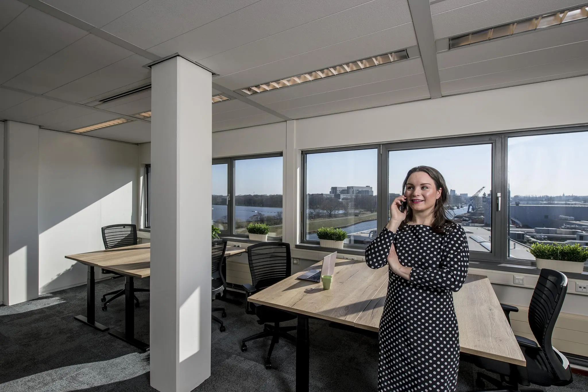 Vrouw in een modern kantoor aan de Vanadiumweg, telefonerend bij een bureau met uitzicht op een industrieterrein.