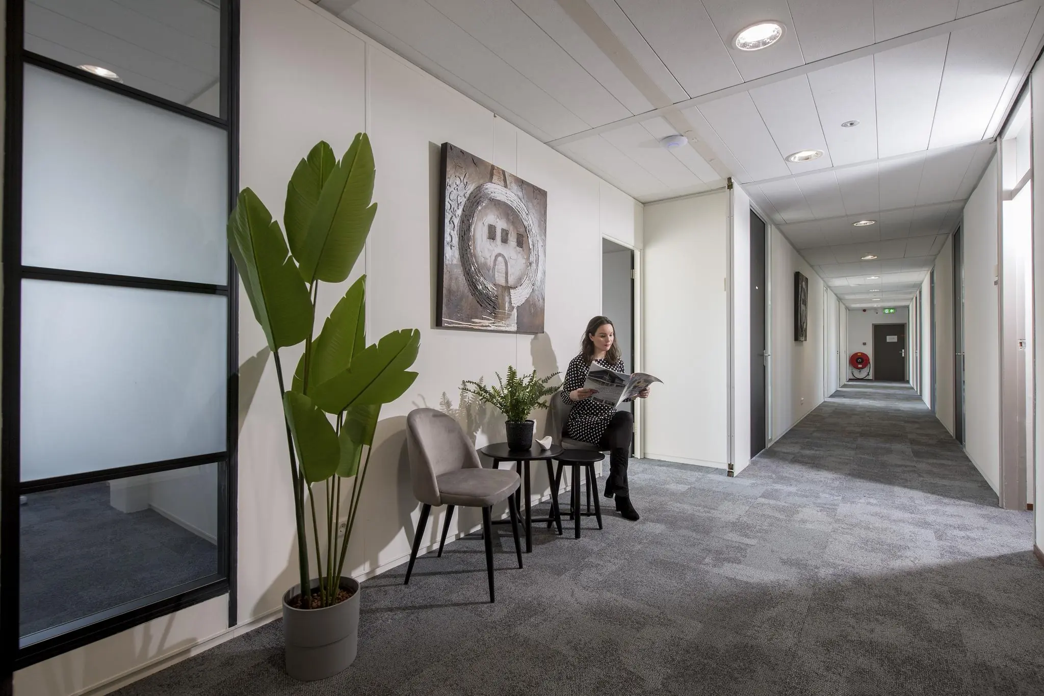 Vrouw leest een tijdschrift in een wachtruimte aan een gang op Vanadiumweg, ingericht met planten en moderne kunst.