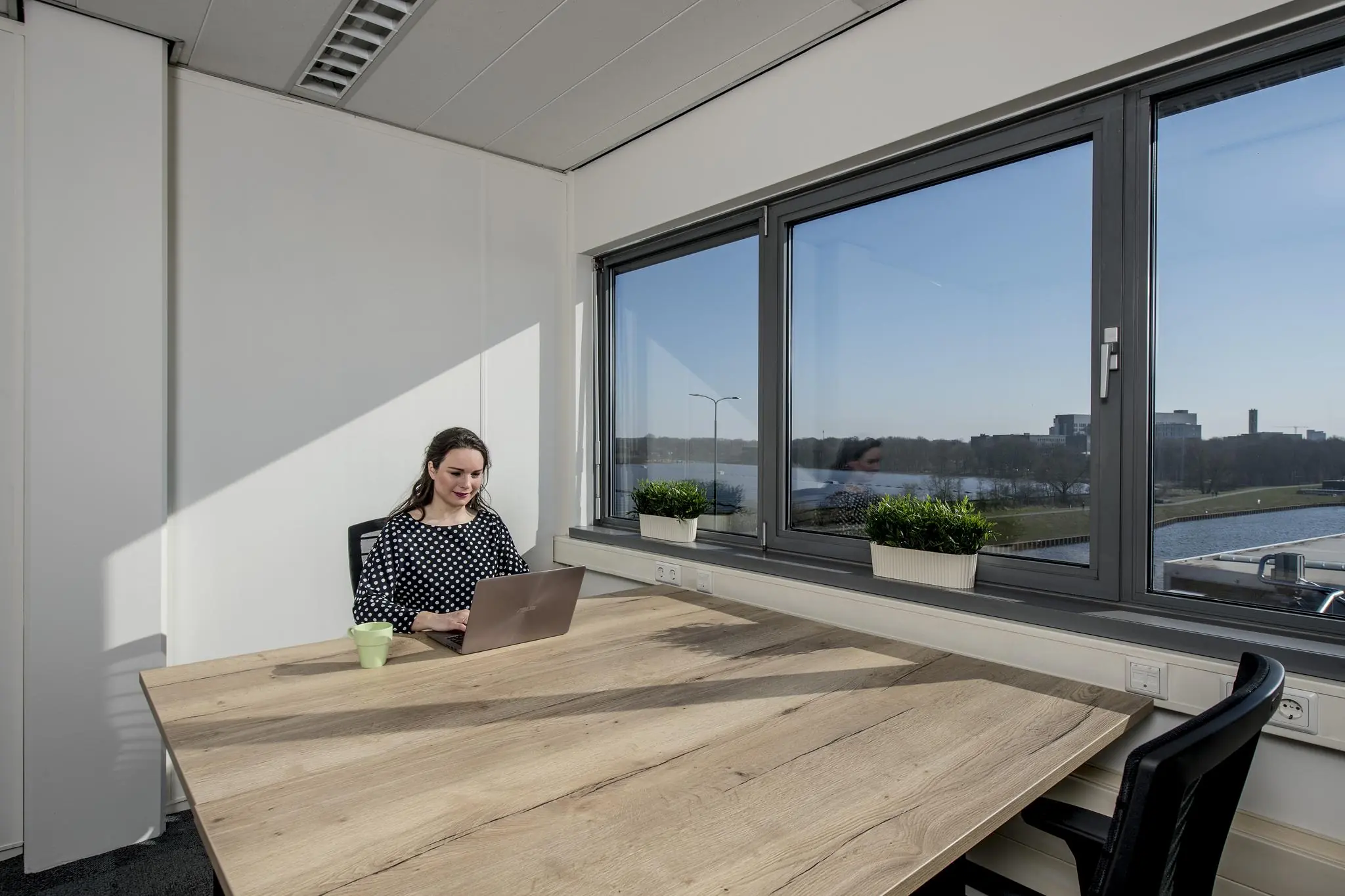 Vrouw werkt op een laptop aan een houten tafel in een zonnig kantoor aan de Vanadiumweg met uitzicht op water en gebouwen.