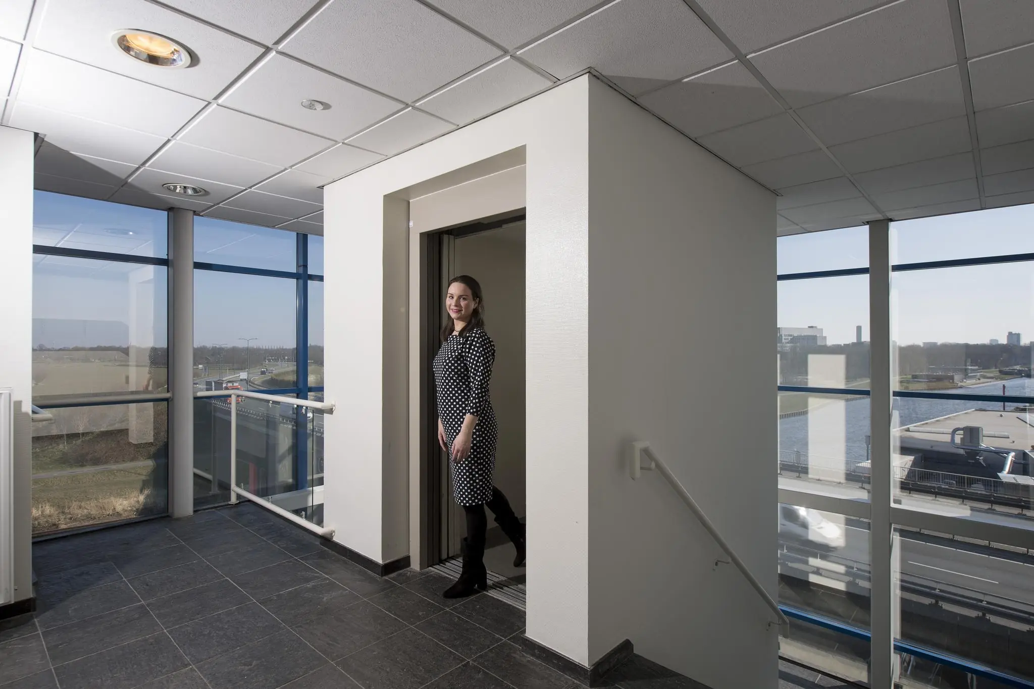 Vrouw stapt lachend uit een lift in een licht en modern kantoorpand aan de Vanadiumweg met uitzicht op weg en water.