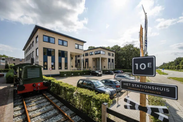 Modern kantoorgebouw aan de Amersfoortseweg met parkeerplaats, oud treinstel en een bord met de tekst "Het Stationshuis – Vergaderen en Ontmoeten".