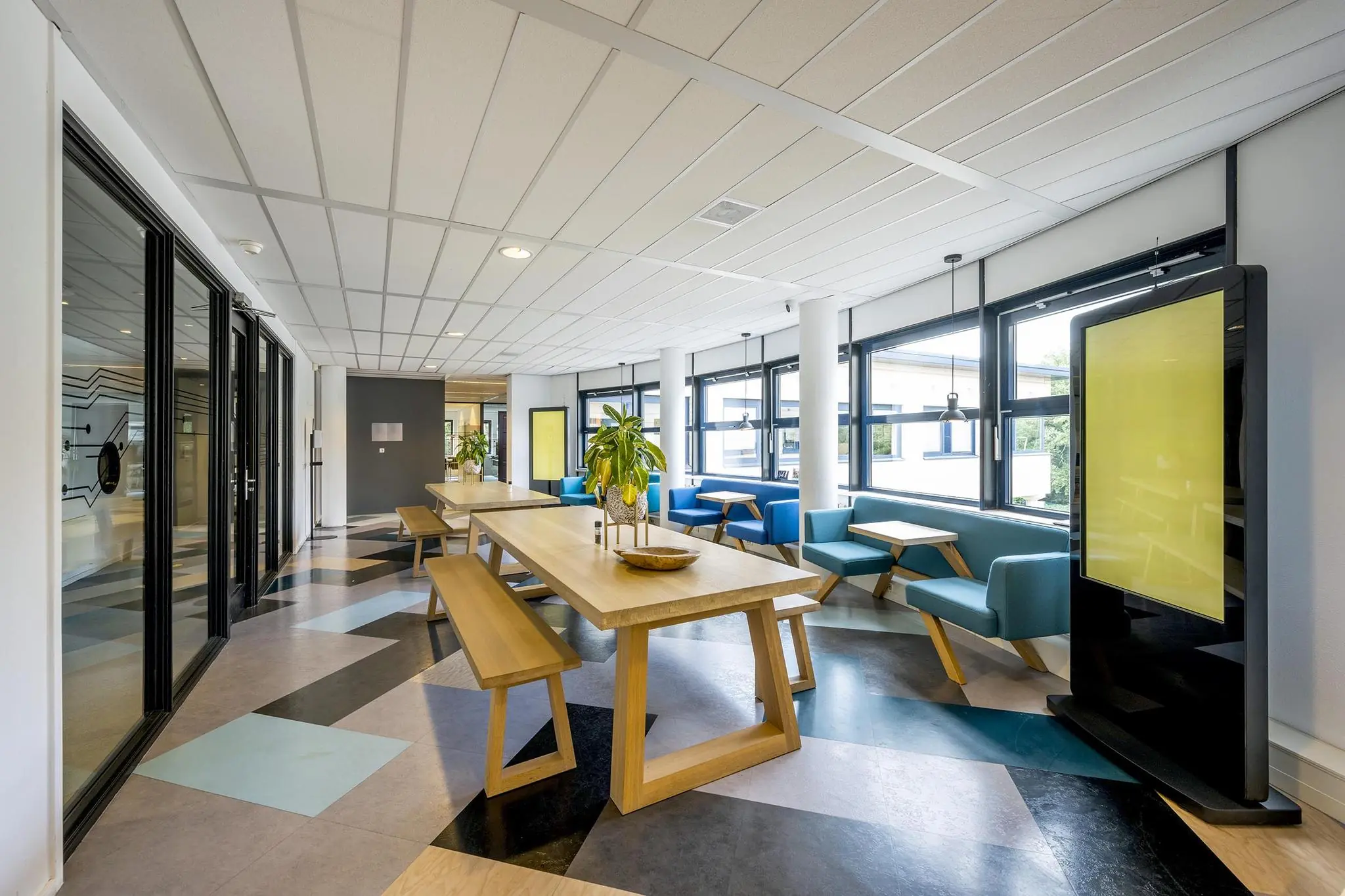 Modern ingerichte ontmoetingsruimte aan de Amersfoortseweg met houten tafels, blauwe zitbanken en veel daglicht.