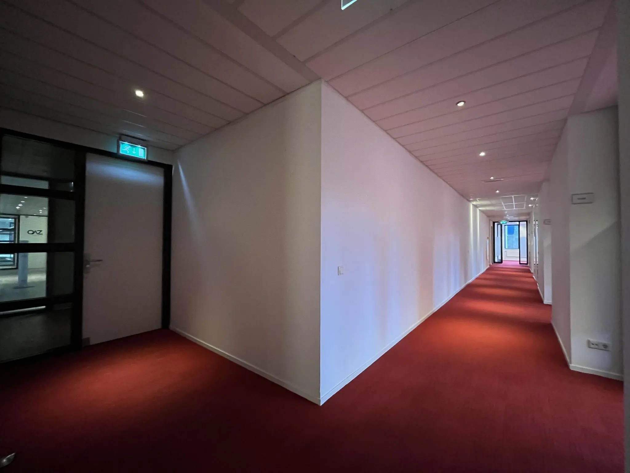 Verlichte gang met rood tapijt in een kantoorpand aan de Amersfoortseweg, met nooduitgangsbord en glazen deur aan het einde.