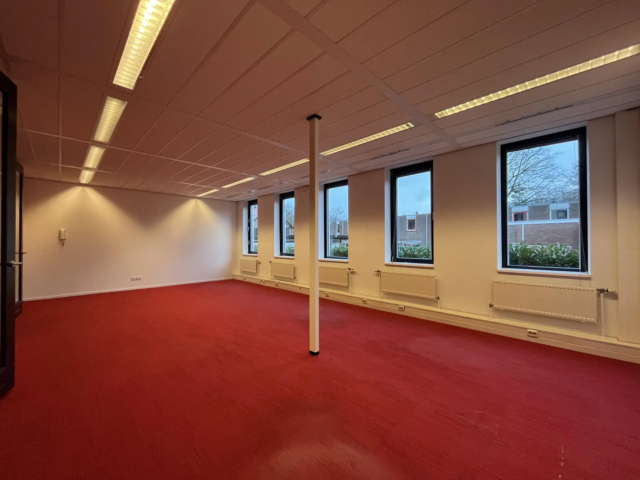 Leeg kantoorruimte aan de Amersfoortseweg met rood tapijt, witte wanden en grote ramen met uitzicht op een woonwijk.