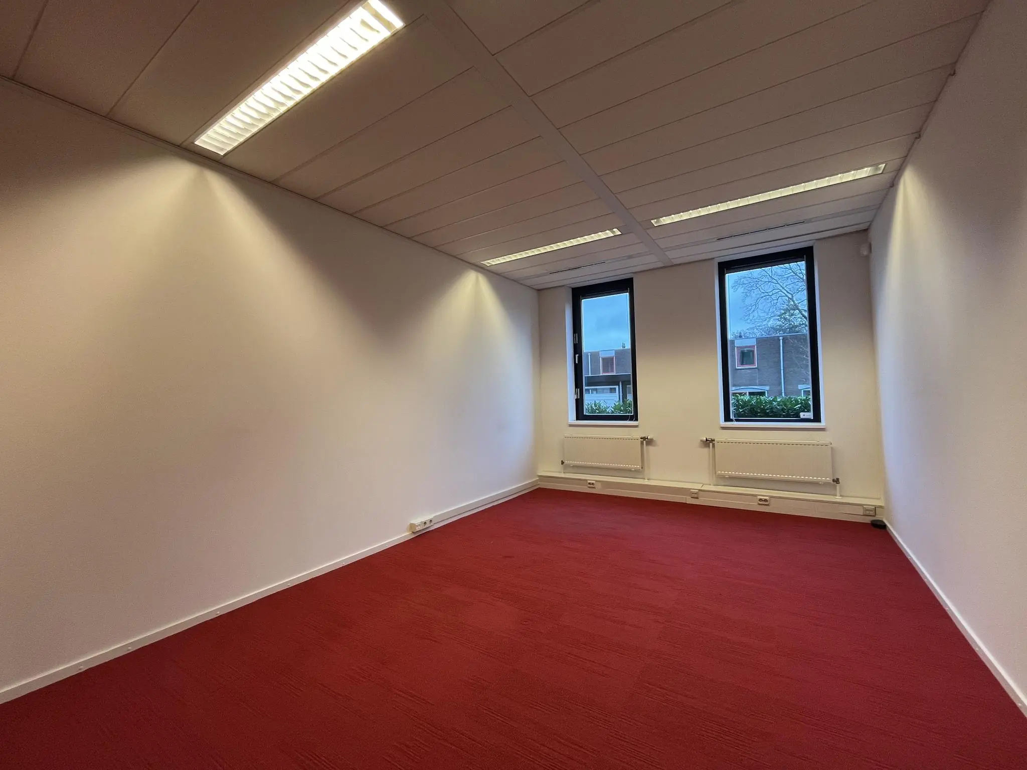 Lege kantoorruimte aan de Amersfoortseweg met rood tapijt, witte muren en twee ramen met uitzicht op een woonwijk.