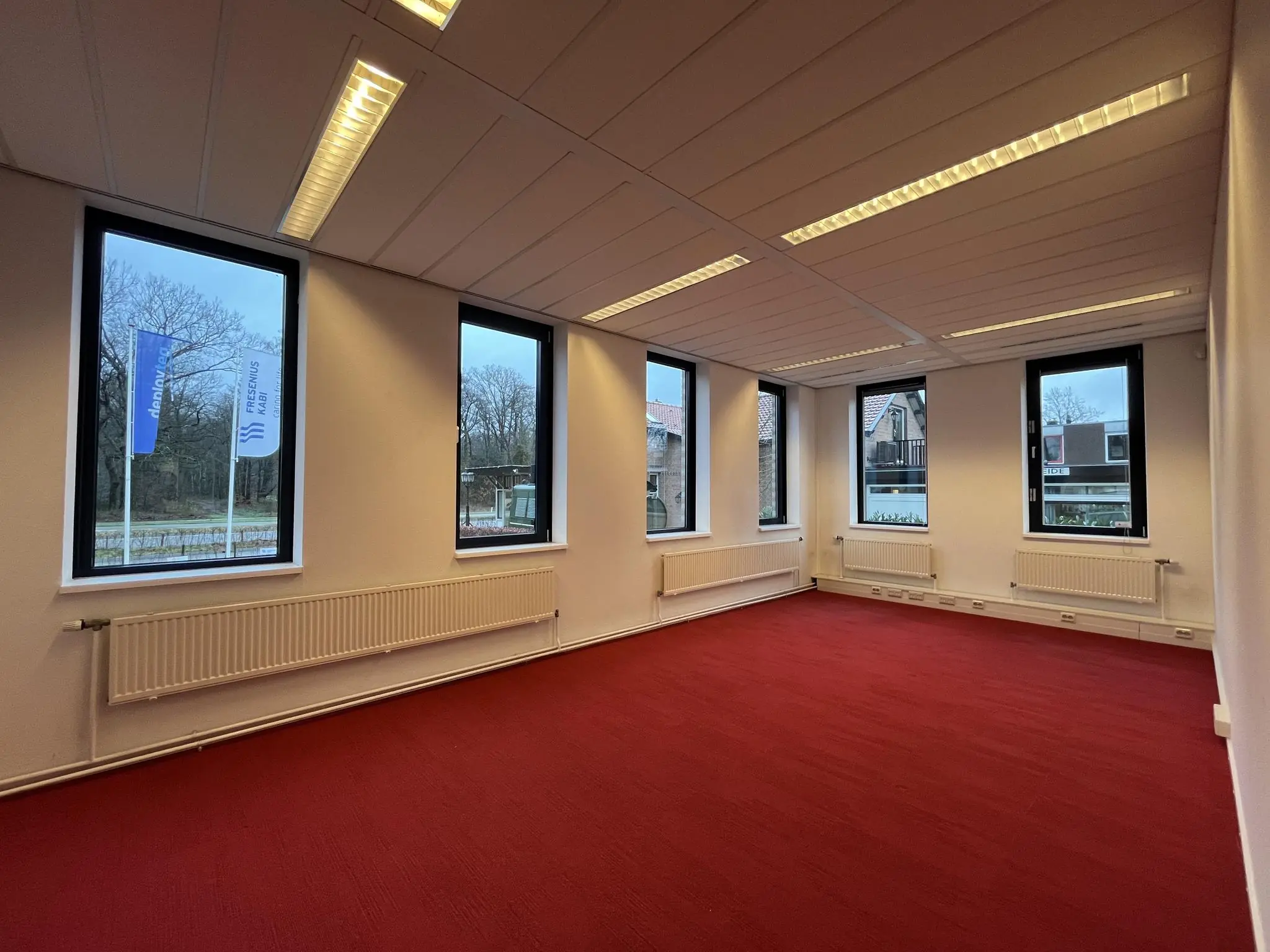Leeg kantoorruimte aan de Amersfoortseweg met rood tapijt, witte muren en meerdere ramen met uitzicht op buiten.