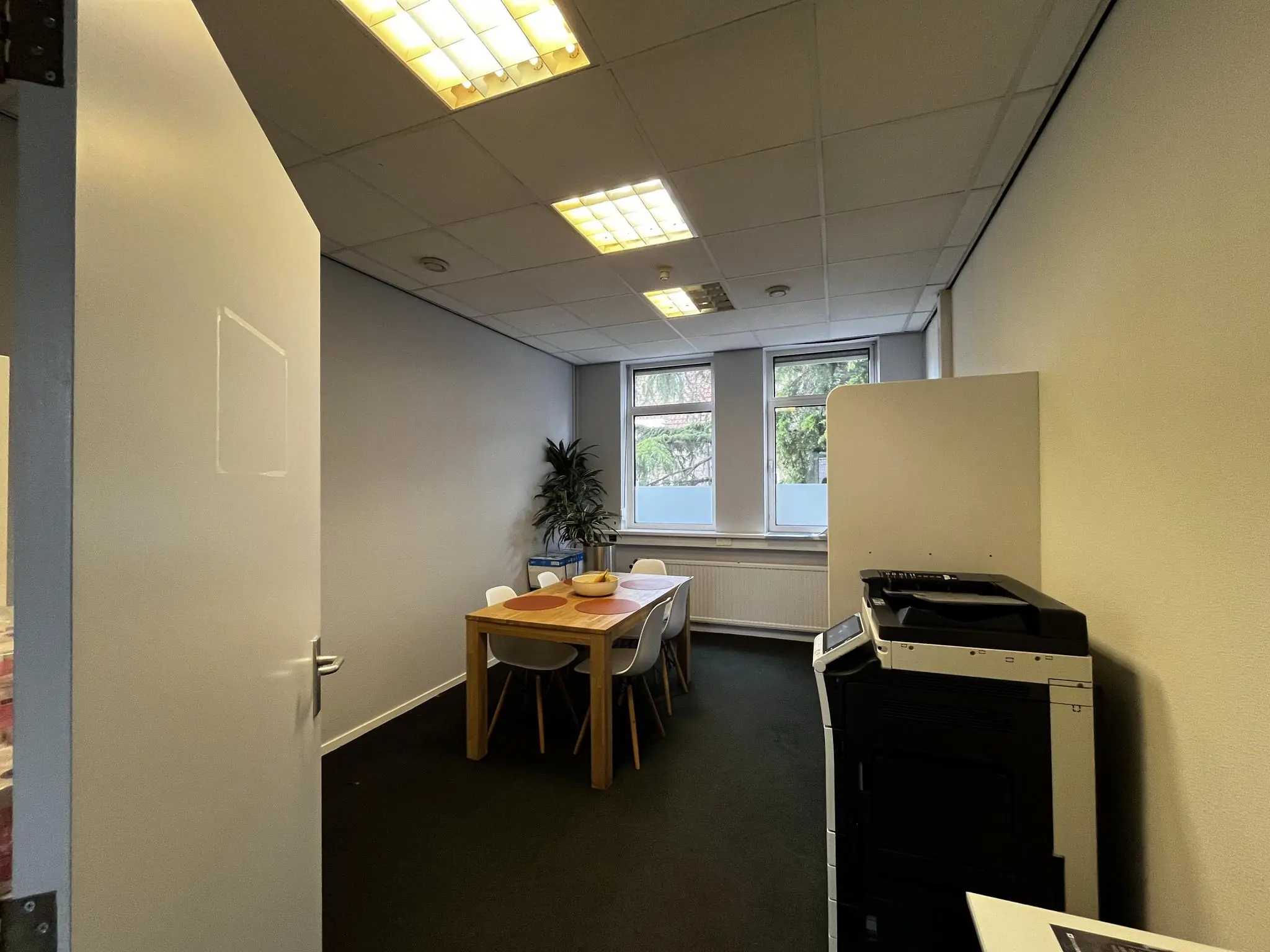 Vergaderruimte aan de Utrechtseweg met een houten tafel, zes stoelen, een printer en drie ramen met uitzicht op groen.