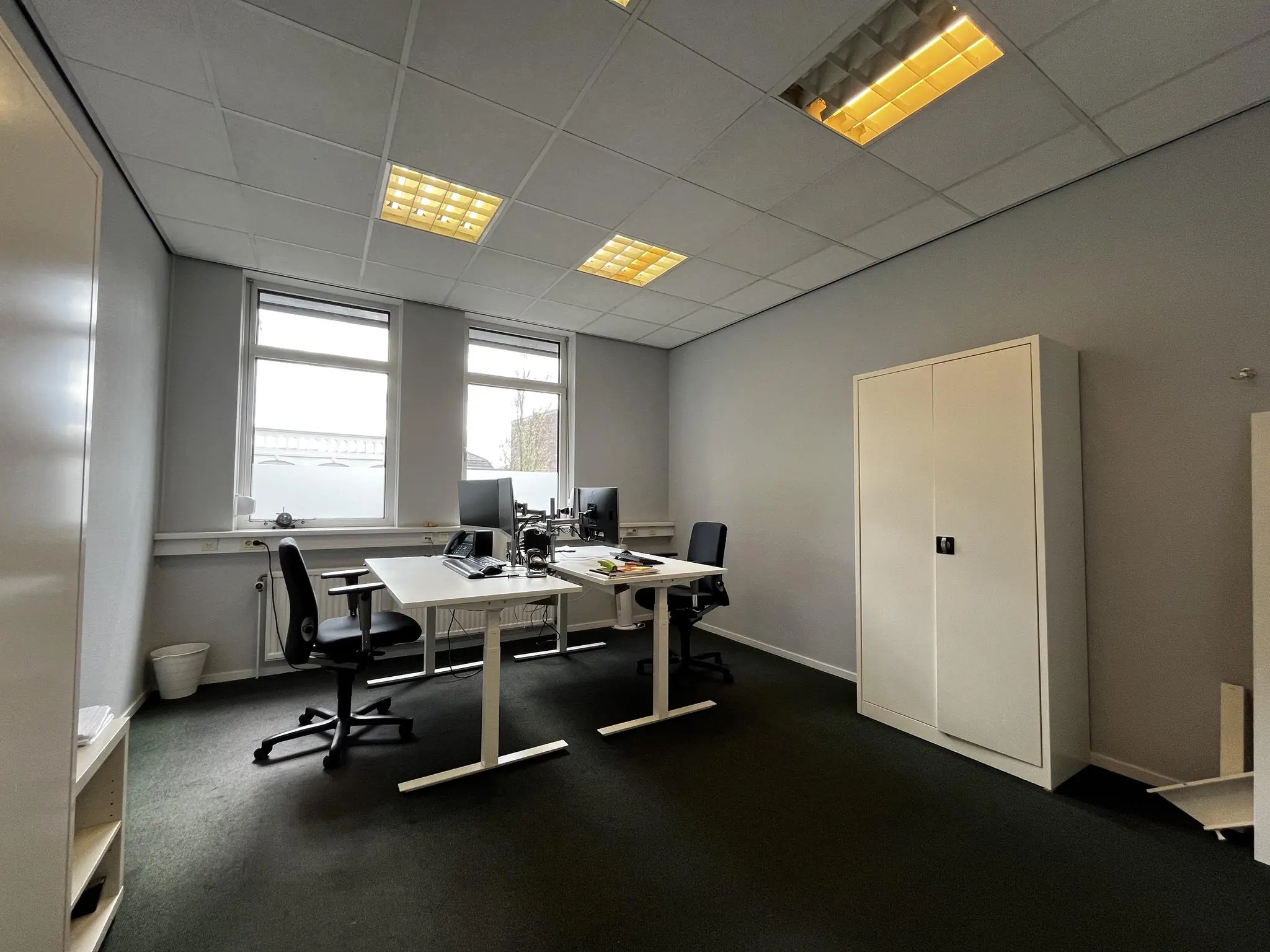 Eenvoudig ingericht kantoor met twee bureaus, bureaustoelen, computers en een opbergkast aan de Utrechtseweg.