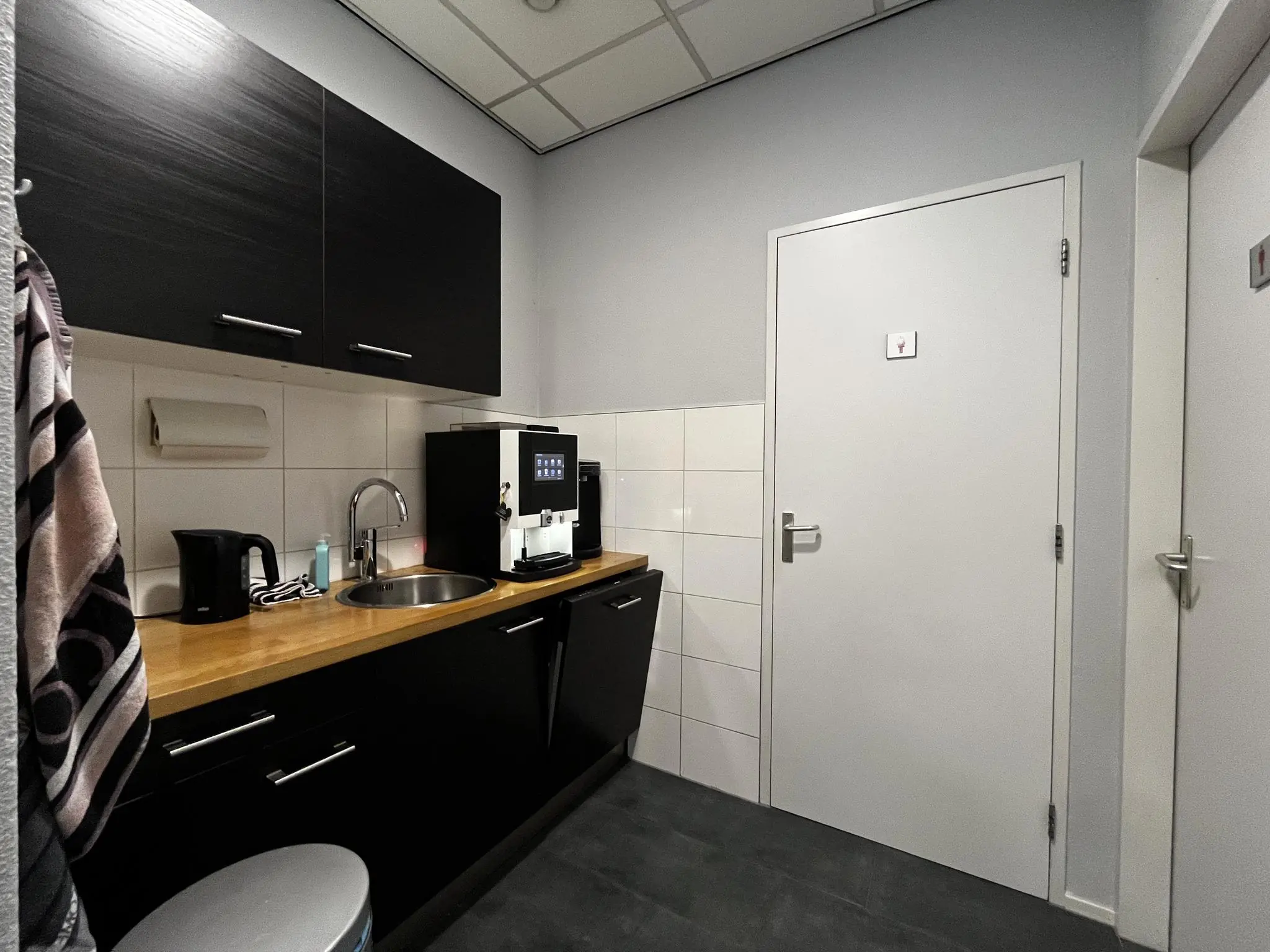 Kleine pantry met zwarte keukenkastjes, een koffieautomaat en een wastafel aan de Utrechtseweg.