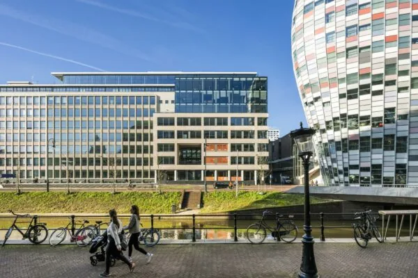 Twee vrouwen lopen met een kinderwagen langs de Catharijnesingel in Utrecht, met moderne kantoorgebouwen en het opvallende bollende gebouw van Hoog Catharijne op de achtergrond.