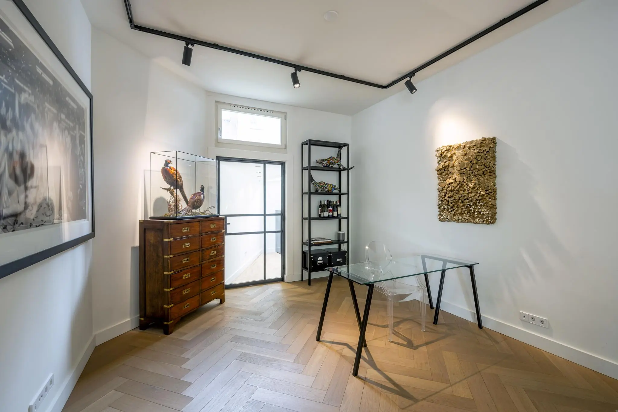Modern ingerichte werkkamer aan de Lomanstraat met glazen bureau, kunstobjecten en houten visgraatvloer.