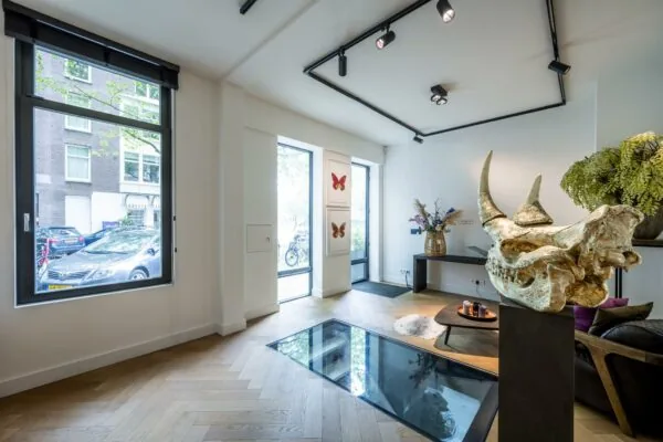 Moderne woonkamer aan de Lomanstraat met een glazen vloerpaneel, kunstobjecten en grote ramen met uitzicht op de straat.