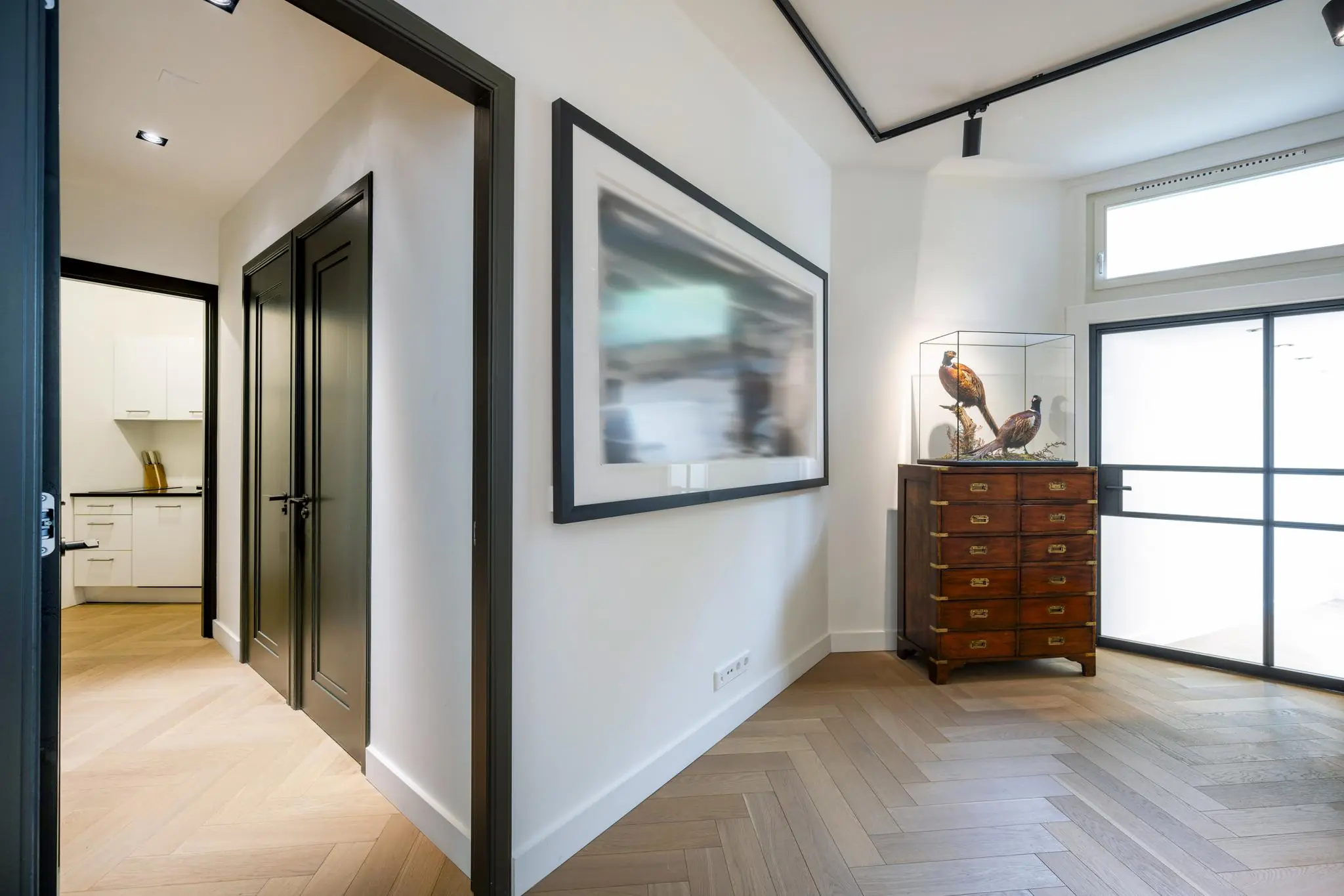 Hal met houten visgraatvloer, een vitrinekast met opgezette fazanten en moderne zwarte binnendeuren in een woning aan de Lomanstraat.