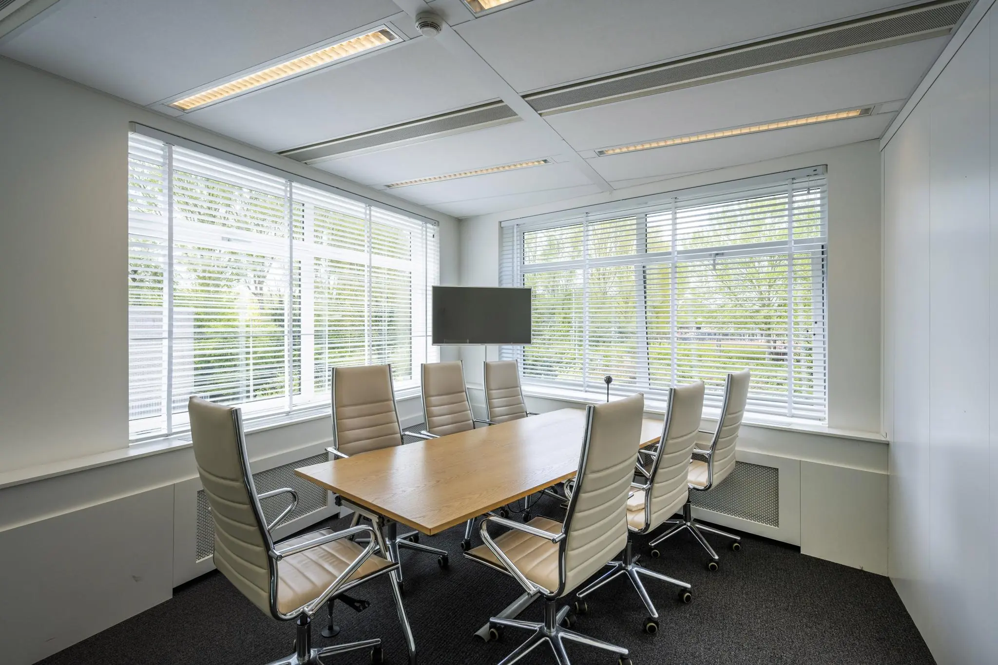 Vergaderruimte aan de Amstelveenseweg met een houten tafel, acht leren stoelen en een flatscreen, omgeven door grote ramen met jaloezieën.