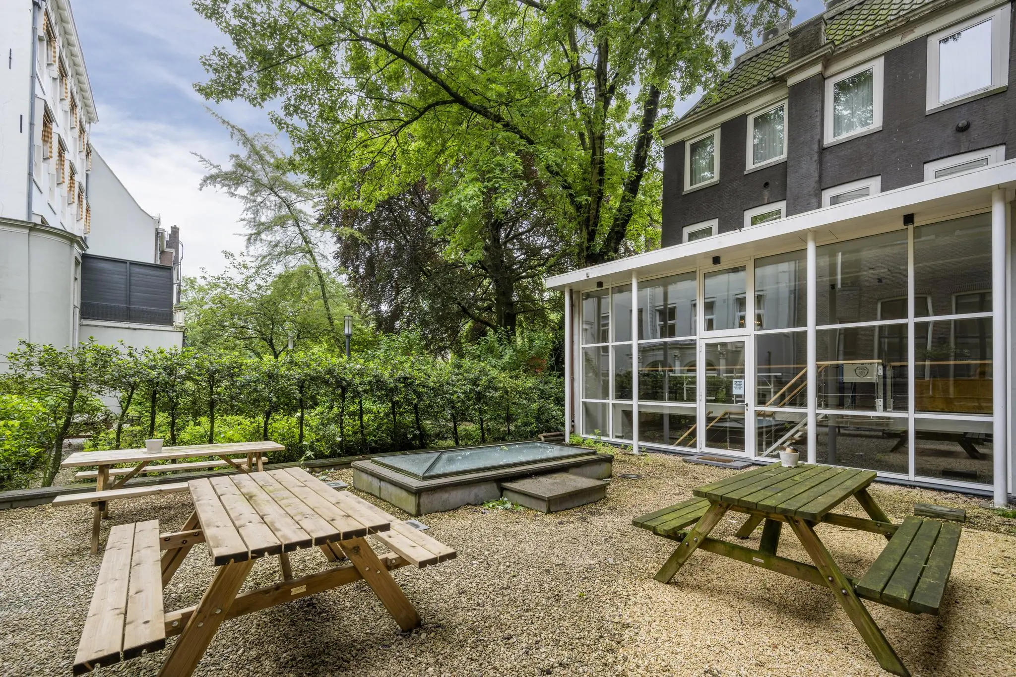 Buitenruimte met picknicktafels naast een glazen gevel van een gebouw aan de Herengracht, omringd door groen.