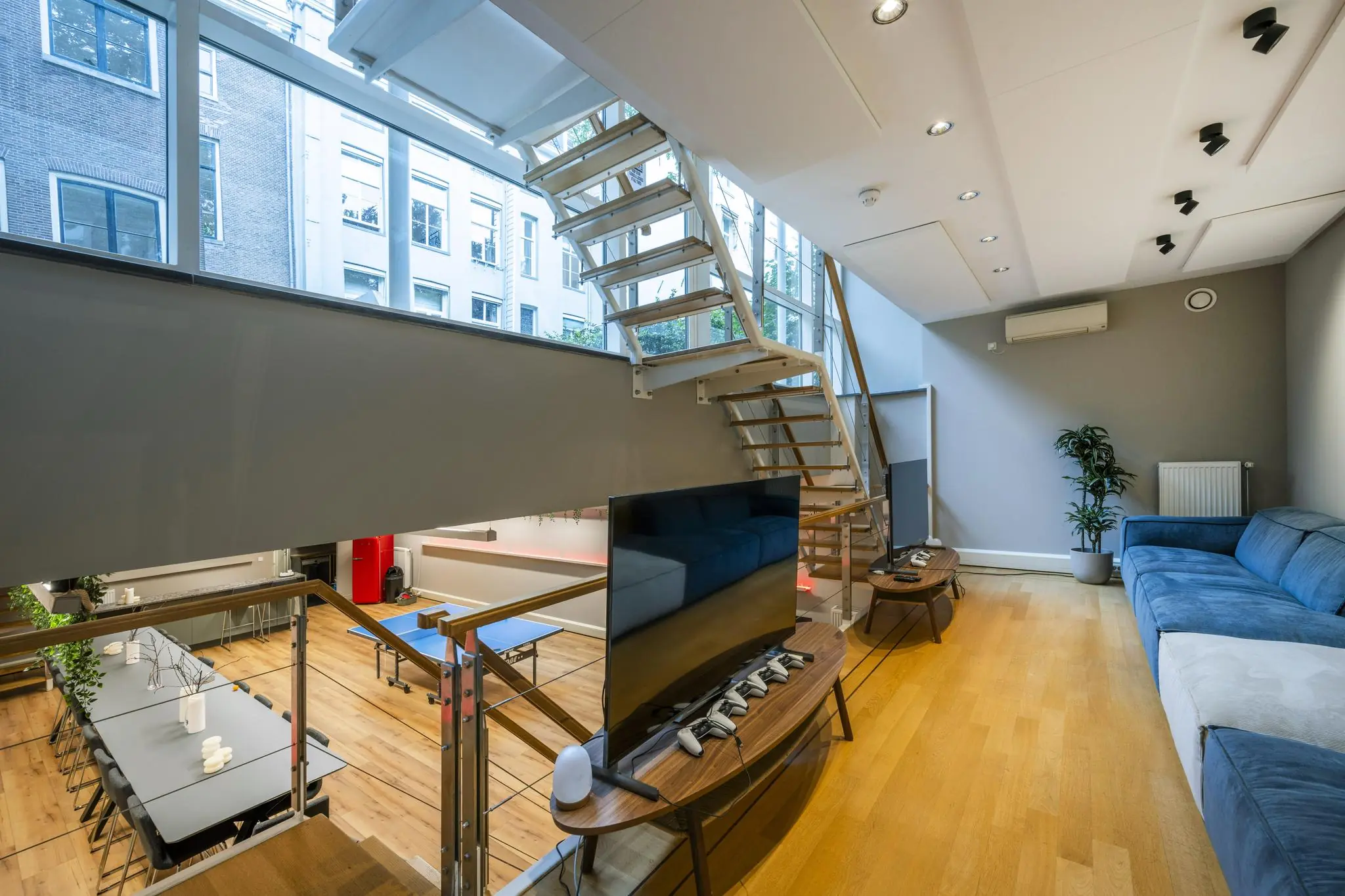 Moderne woonkamer aan de Herengracht met een speelse split-level indeling, grote ramen, een blauwe bank en een zicht op een pingpongtafel beneden.