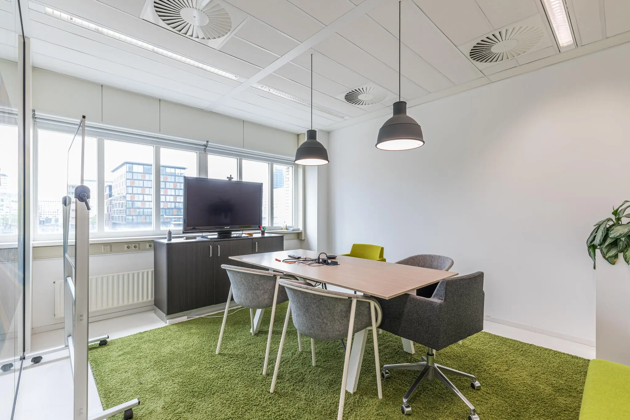 Moderne vergaderruimte in kantoorgebouw Leidseveer met grote ramen, groen vloerkleed en een flatscreen op kast.