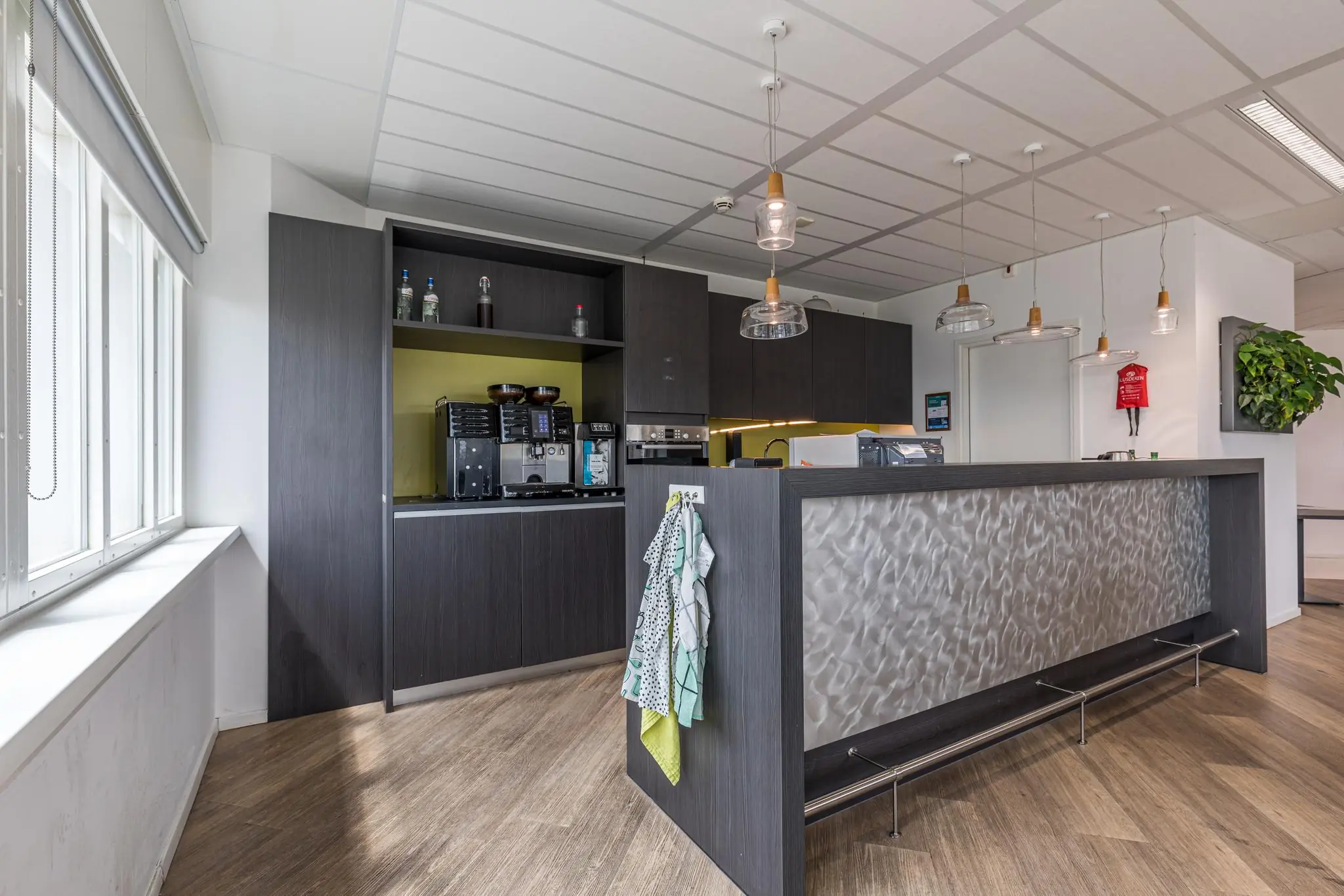 Moderne kantoorpantry met koffieautomaat, hanglampen en een balie met roestvrijstalen afwerking in het Leidseveer-gebouw.